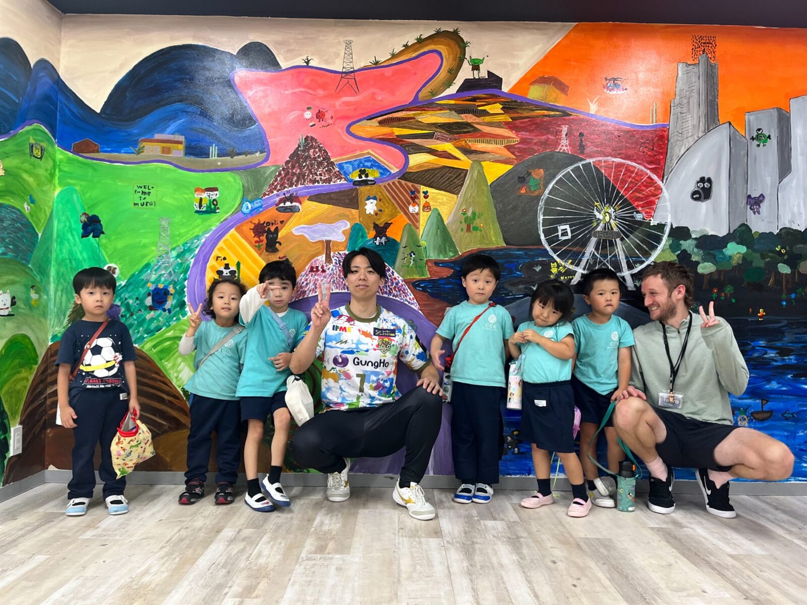 Seiya Kitano, teachers and children posing together in front of CGK's mural